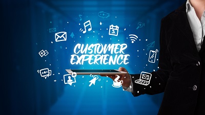 A person in a suit holding a tablet with the phrase 'CUSTOMER EXPERIENCE' displayed above it, surrounded by icons including an envelope, music note, shopping cart, thumbs up, coffee cup, laptop, and Wi-Fi signal, set against a blue digital-themed background.