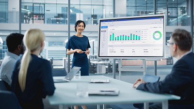 A business meeting with four people, where one person is standing and presenting data on a large screen displaying bar charts and a pie chart, while three others are seated at a table observing the presentation.